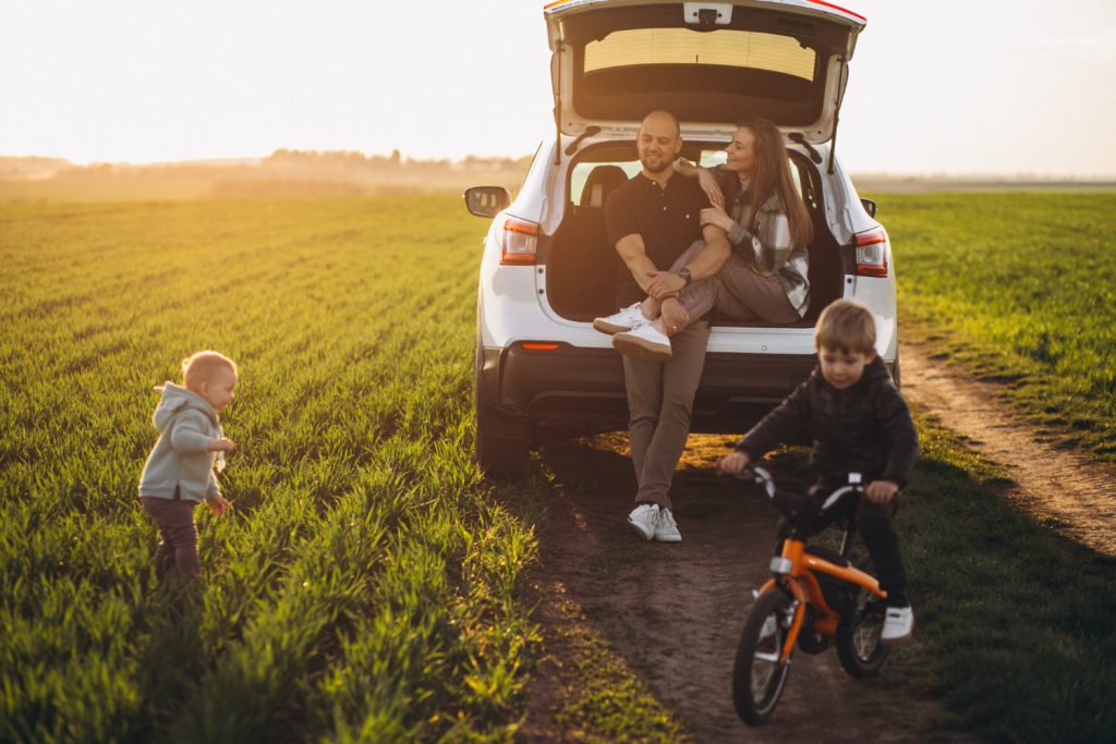 Jeune famille charge des valises dans le coffre d'un SUV moderne sur l'allée d'une maison de banlieue au matin. Les parents et deux enfants portent des vêtements décontractés, la lumière douce et volumétrique crée une ambiance naturelle et détendue.